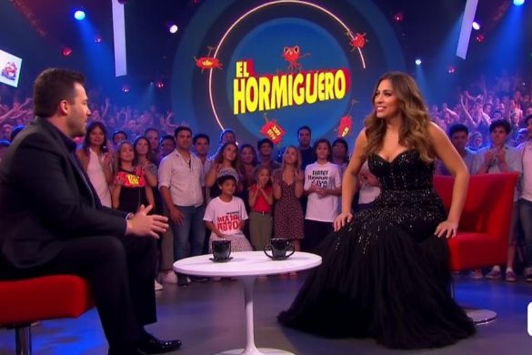 La escena está ambientada en un vibrante estudio de televisión lleno de luces coloridas y una audiencia animada. En el centro, la carismática cantante 'Mónica Naranjo' está sentada en una elegante silla moderna, con un elegante vestido negro adornado con lentejuelas brillantes que captan la luz, enfatizando su presencia dinámica. Su cabello está peinado en olas glamorosas, enmarcando su rostro, que está iluminada con una cálida sonrisa mientras entabla una conversación con el anfitrión 'Pablo Motos', que se inclina animadamente hacia adelante, mostrando su entusiasmo. El telón de fondo presenta el logotipo de la programa 'El Hormiguero' en letras audaces y juguetones, rodeadas de decoraciones caprichosas, incluidos los icónicos 'Formigues' o hormigas, 'Trancas' y 'Barrancas', que son dibujos animados y cómicamente de gran tamaño, agregando un toque de humor a la atmósfera. La audiencia está cautivada, con algunos fanáticos que sostienen señales que leen 'No Me Retiro' y 'Requiro Temps', lo que refleja su apoy