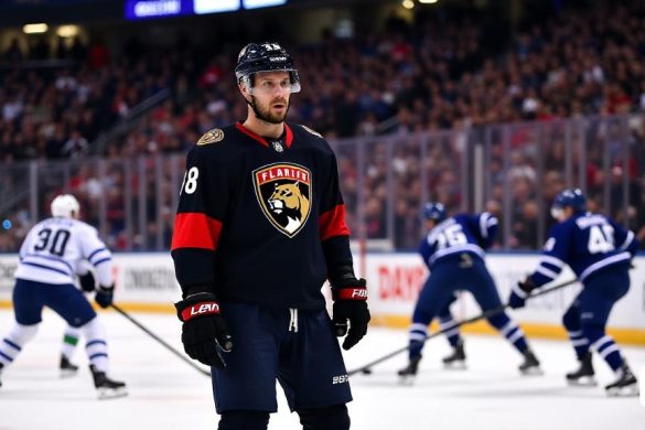 La imagen captura un momento tenso en un juego de hockey profesional, mostrando la intensidad del hockey de playoffs. En primer plano, Aaron Ekblad, un defensa alto y musculoso de los Florida Panthers, se ve en su camiseta del equipo negro y rojo, con el logotipo de 'Panthers' que se exhibe prominentemente en su pecho. Su expresión está enfocada y determinada, lo que refleja la presión del juego. Detrás de él, la pista de hielo está llena de acción; Los jugadores de ambos equipos se dedican a una feroz batalla por el disco. Los Toronto Maple Leafs, vestidos con sus clásicos uniformes azules y blancos, parecen estar a la defensiva, con un jugador intentando bloquear un disparo mientras otro patina difícil de recuperar la posesión. La multitud en las gradas es un borrón de emoción, con los fanáticos agitando banderas y con colores de equipo, creando una atmósfera vibrante. El marcador en el fondo muestra una puntuación cerrada, aumentando la tensión del momento. La iluminación es brillante, iluminando el hielo