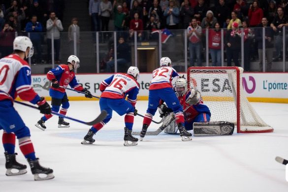 Una escena dinámica e intensa de un partido de hockey sobre rolleros entre 'El Club Patí Voltrregà' y 'Ch Caldes Recam Làser'. La imagen captura el momento en que 'Arnau Canal' obtiene un objetivo poderoso, enviando el balón a la red, con el marcador mostrando un tenso '2-0' a favor del equipo Voltregà. Los jugadores están en sus sorprendentes uniformes 'Blanc-i-Blau', mostrando su determinación y enfoque. En el fondo, la multitud está de pie, una mezcla de emoción y anticipación, con algunos fanáticos agitando banderas y otros capturando el momento en sus teléfonos. La pista está bien iluminada, destacando los movimientos rápidos de los jugadores y los colores vibrantes de su equipo. La atmósfera es eléctrica, llena de los sonidos de los patines en la pista y los vítores de los seguidores, a medida que el juego progresa a una fase crucial, con ambos equipos compitiendo por un lugar en la próxima competencia europea.